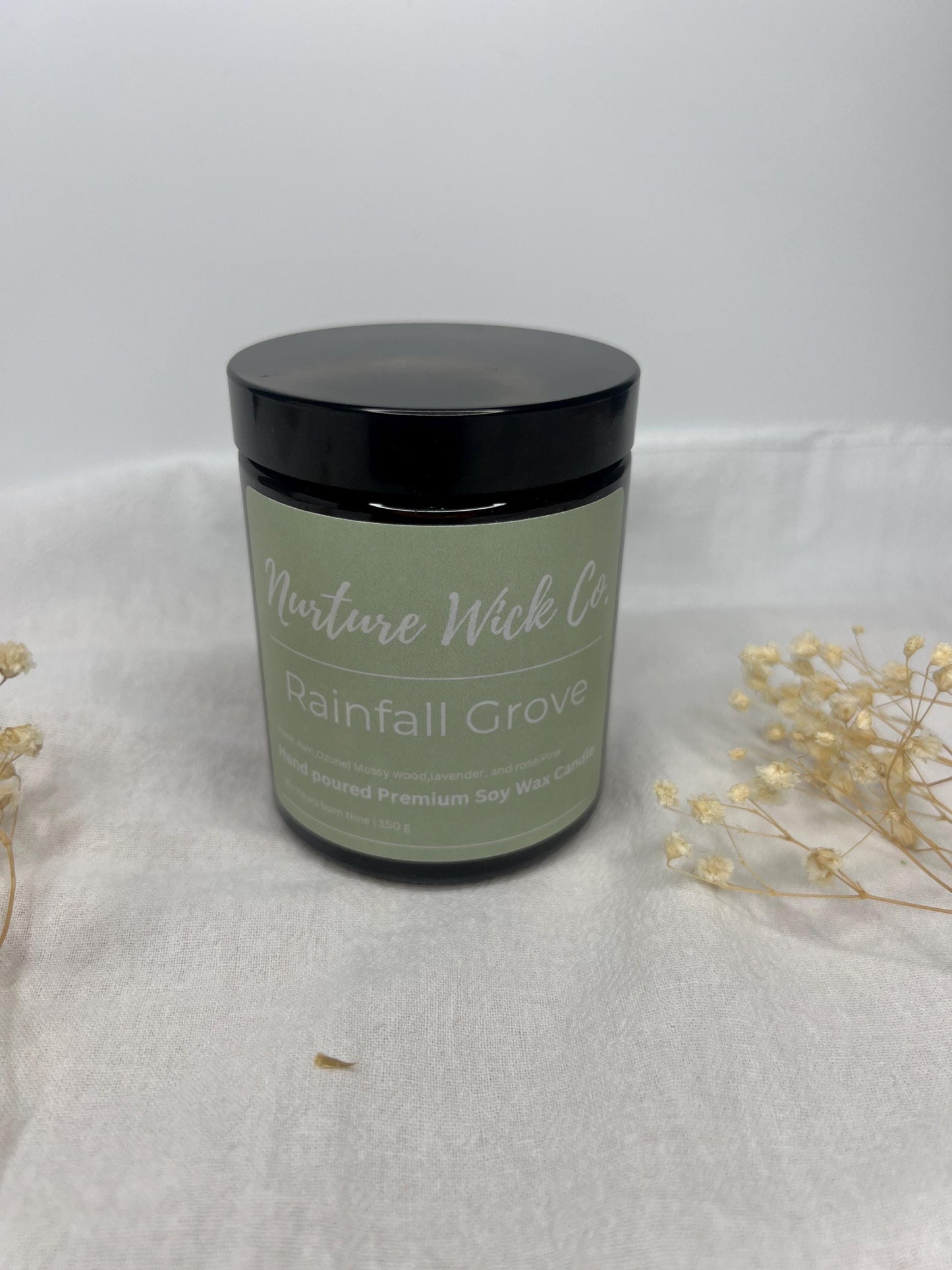 Candle jar labeled 'Rainfall Grove' by 'Nurture Wick Co.' on a white background with dried flowers.