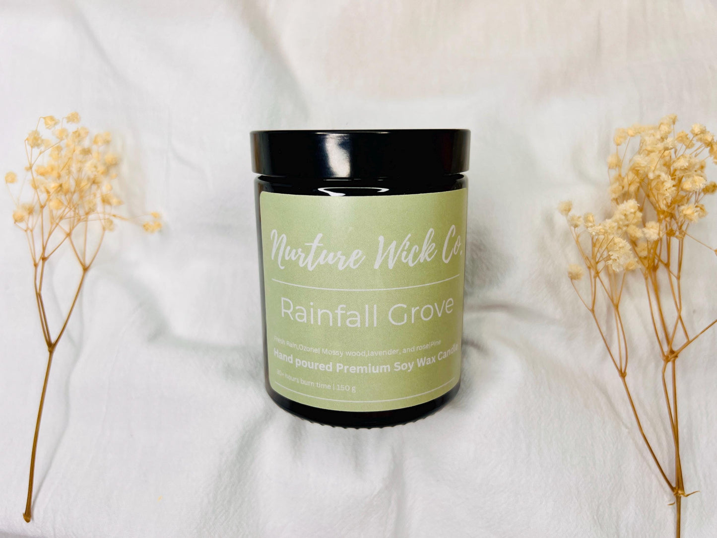 Candle jar labeled 'Rainfall Grove' from Nature Wick Co. on a white fabric background with dried flowers.