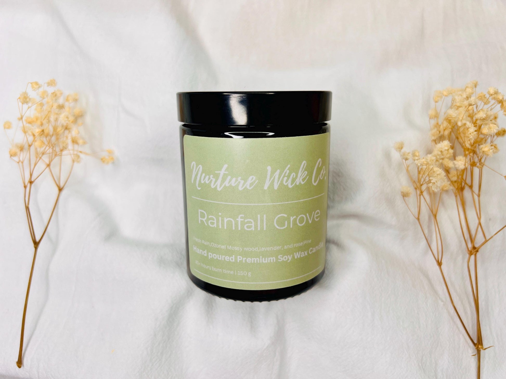 Candle jar labeled 'Rainfall Grove' from Nature Wick Co. on a white fabric background with dried flowers.