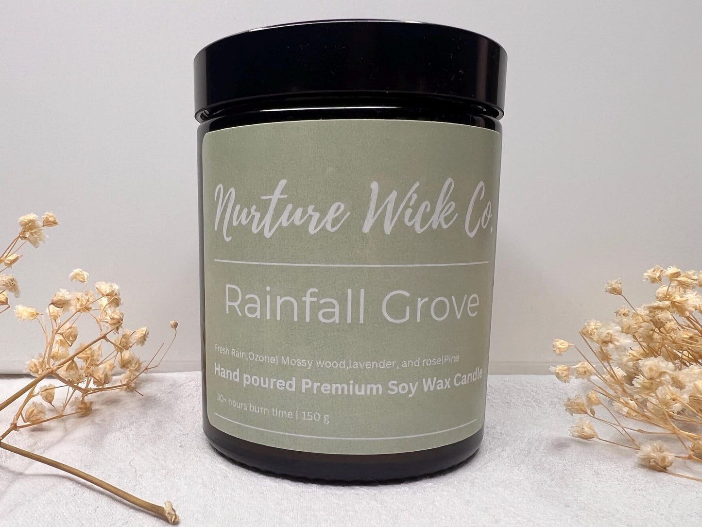 Candle jar labeled 'Rainfall Grove' from Nurture Wick Co on a white surface with dried plants.