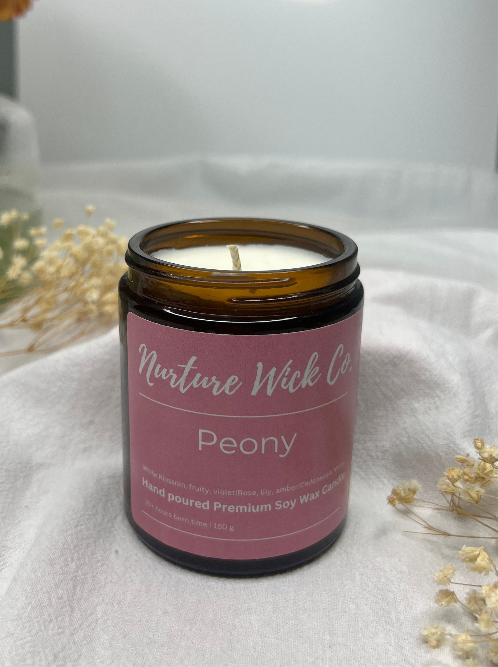 Candle in a glass jar with a pink label on a white surface, surrounded by flowers.